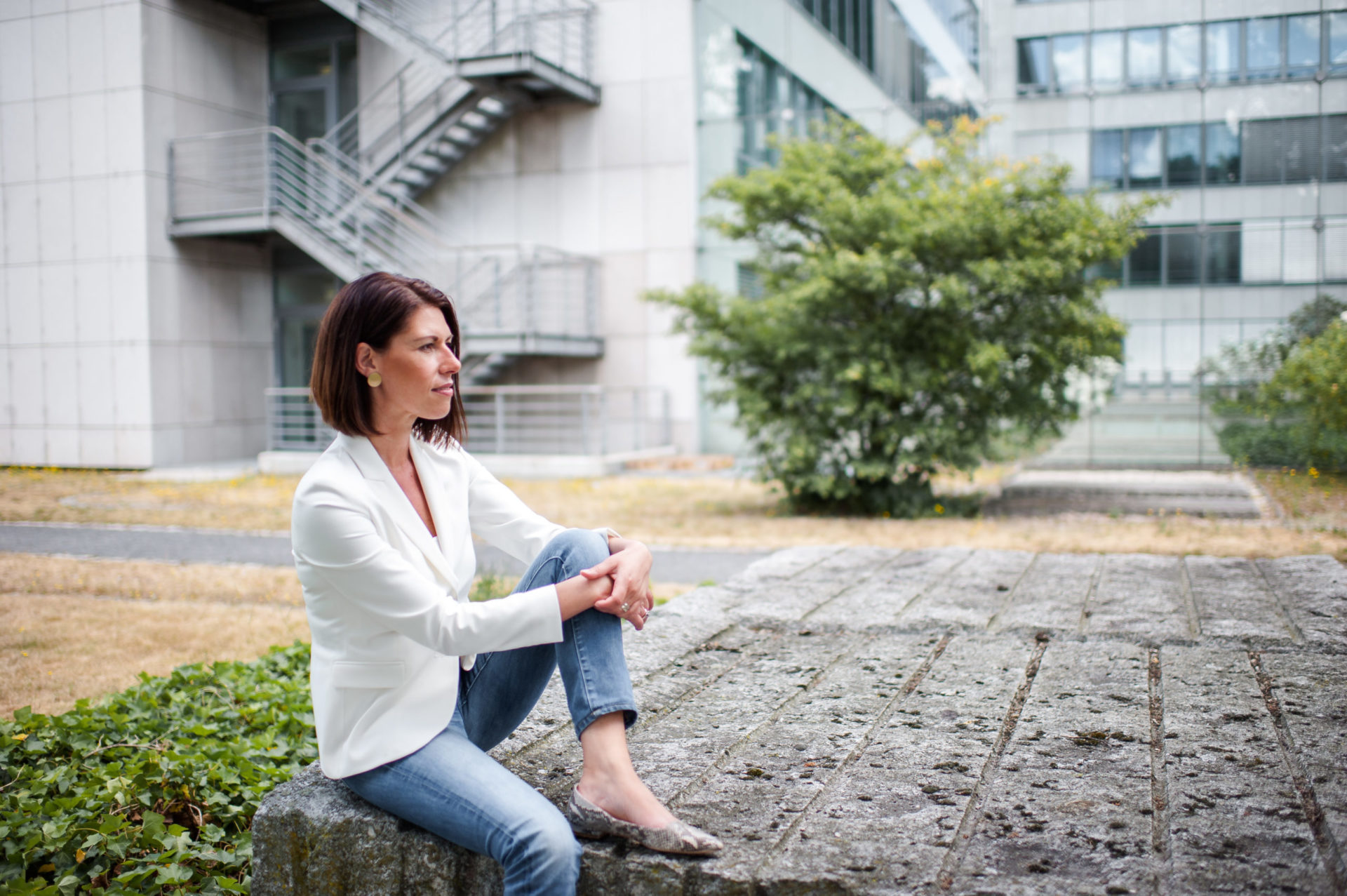 Andrea Lawlor sitzt vor einem Bürogebäude auf einem Stein und blickt in die Ferne